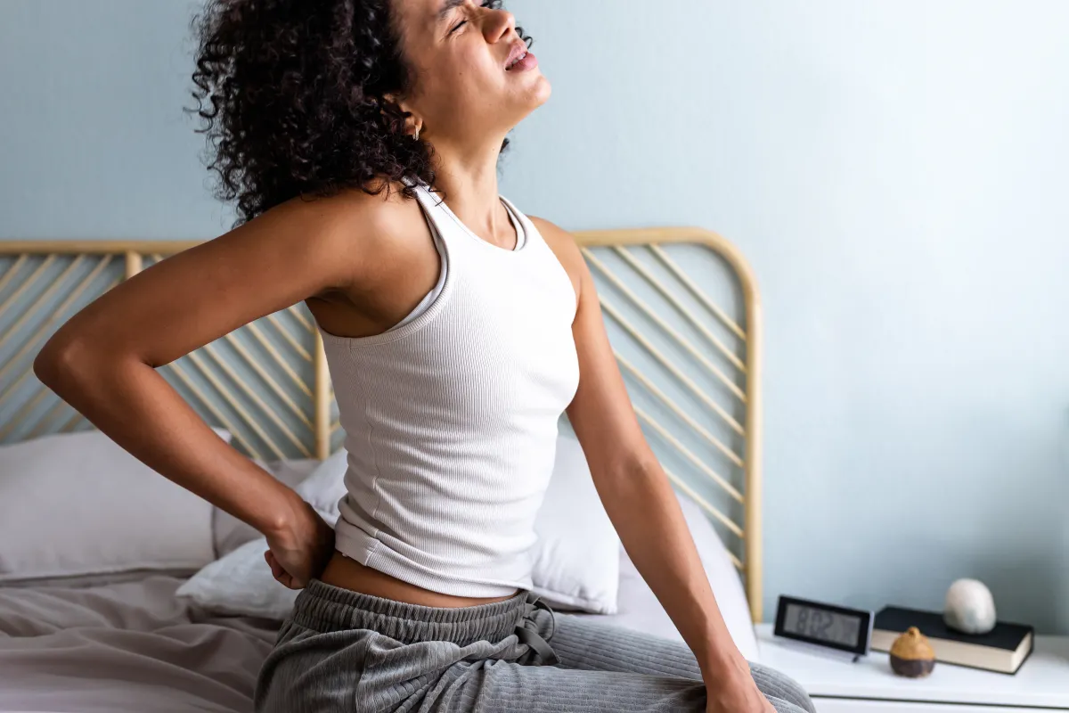 Person sitting on the edge of a bed holding their lower back in pain after waking up