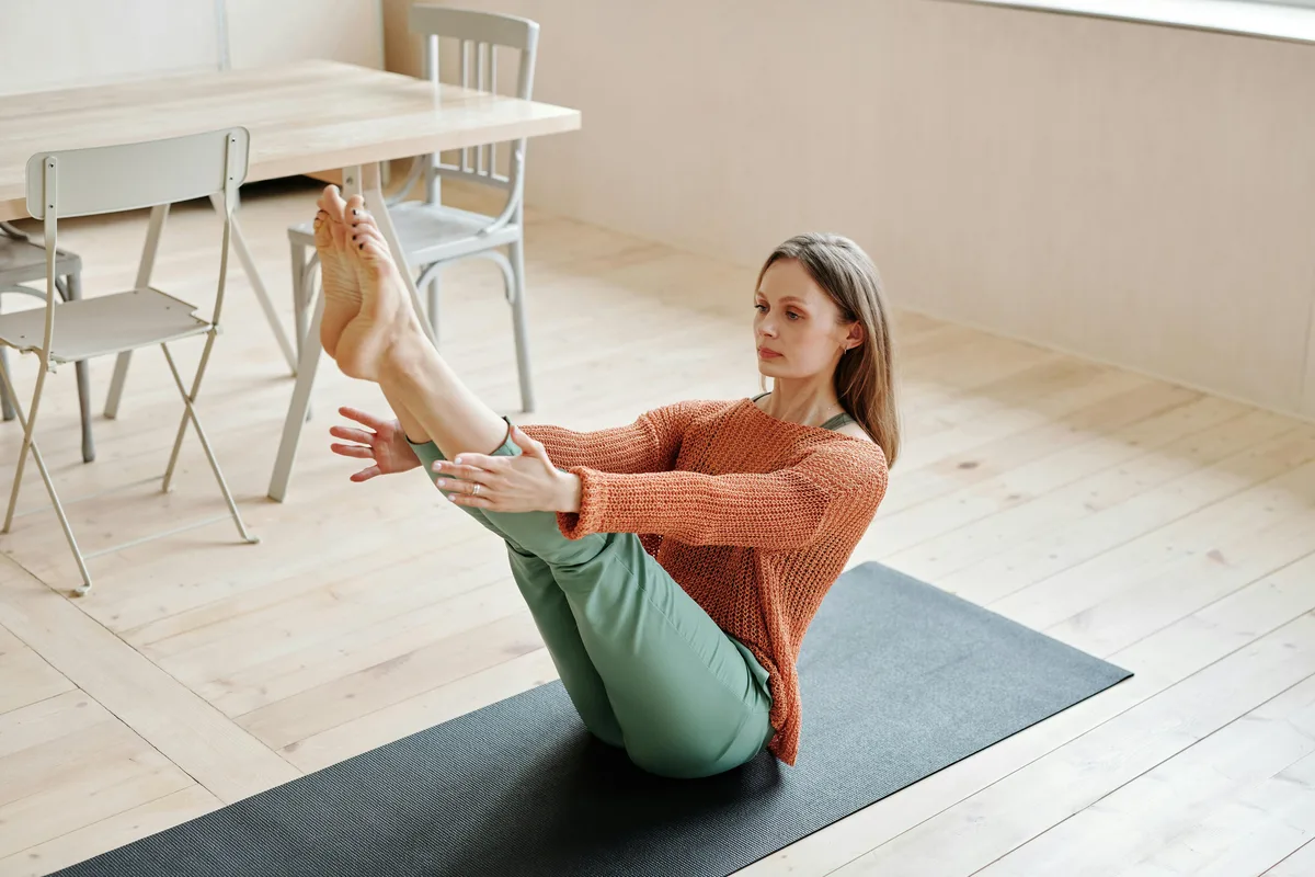 Woman performing a Pilates teaser, a seated core exercise, on a mat at home